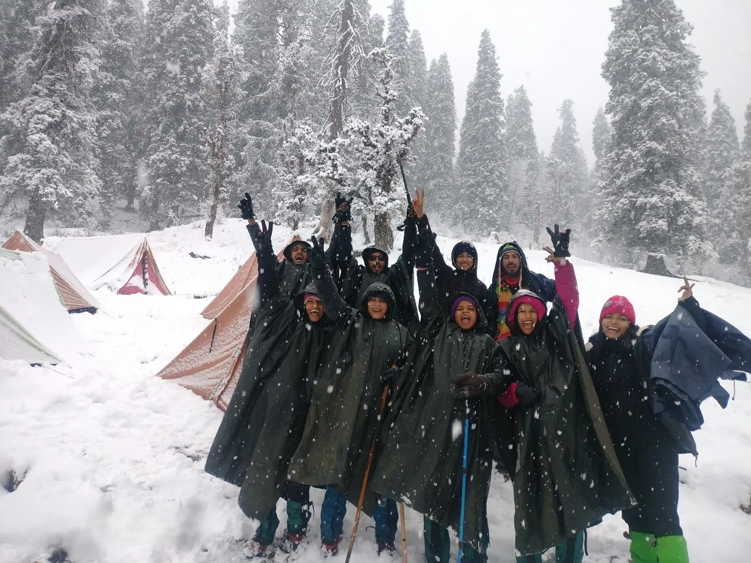 a group of happy trekkers standing in a snow covered campsite on the kedarkantha trek